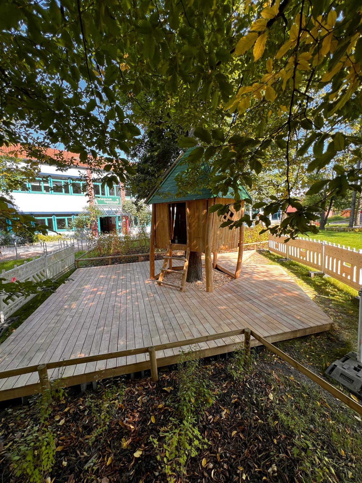 Baumhaus aus Holz mit grünem Dach, um einen Baum gebaut, mit großem umlaufendem Holzdeck